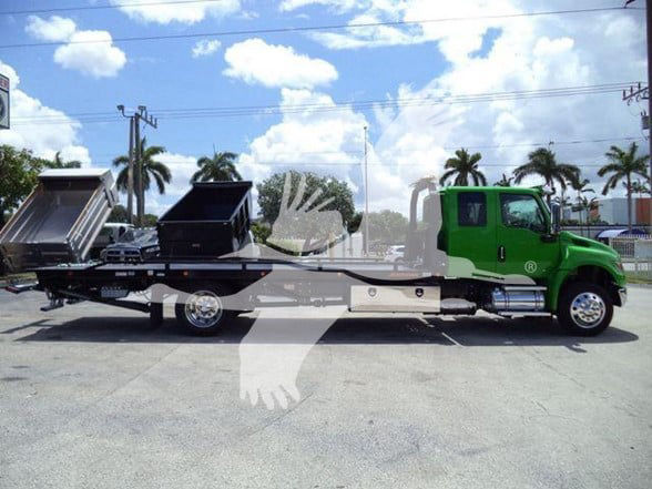 International MV tow truck with green cab and flatbed, loading two black dump containers on a trailer.
