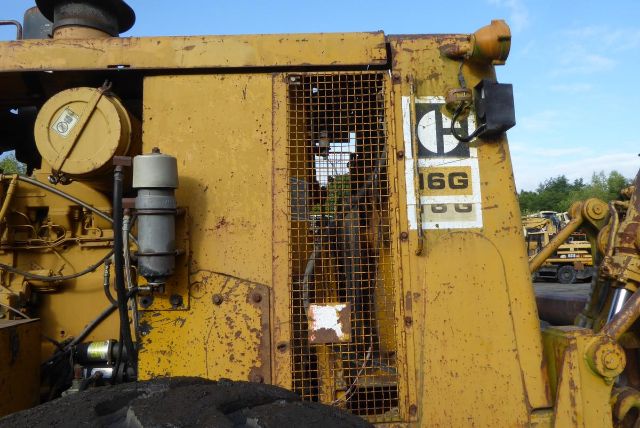 Caterpillar 16G grader cab side showing coolant reservoir and mesh grille with 16G label.