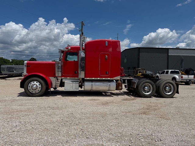 Red Peterbilt 379 conventional sleeper truck in a gravel lot with a black metal building in the background.