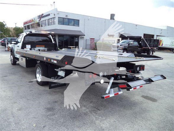 Ford F-550 tow truck with flatbed in service lot, rear view showing hydraulic bed and ramps.