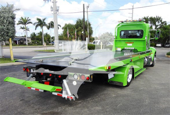 Green 2022 Peterbilt 337 tow truck with flatbed in a lot, rear view shows platform and towing apparatus.