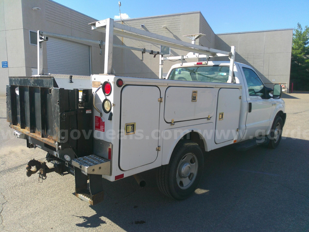 Pickup truck utility body with storage compartments showing side doors and rear black utility bed; visible Ford F-Series style cab; ladder/utility racks on top rear; tow hitch and step.