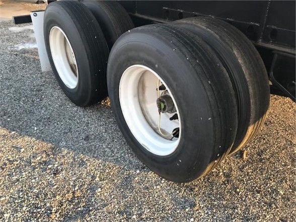 Flatbed trailer dual-axle wheel assembly with black tires and white rims, visible undercarriage and gravel surface.