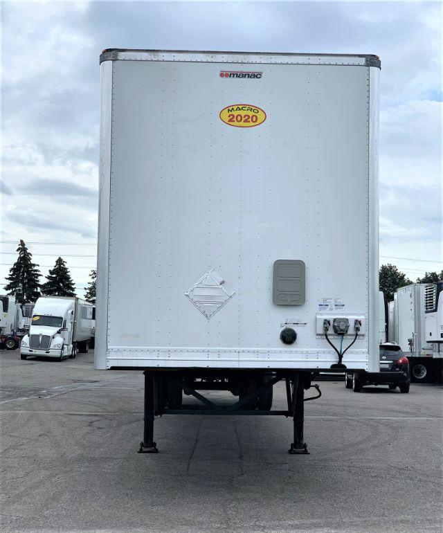 Manac dry van trailer rear view showing bare concrete landing gear and electrical/connectors. MACRO 2020 sticker on door, diamond hazard placard, grey access panel.