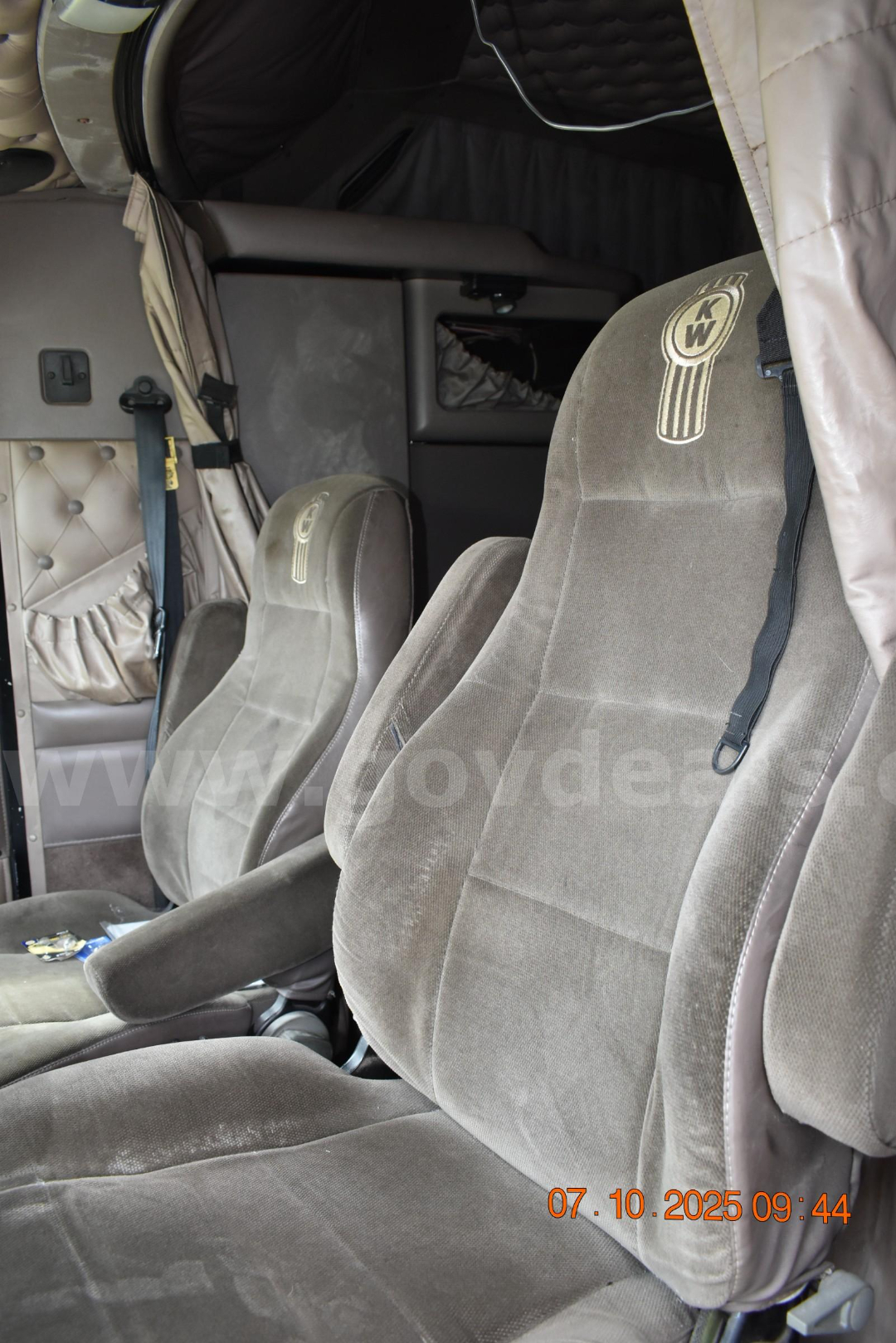 Cab interior of a 2003 Kenworth W900 showing driver and passenger seats with KW logos, beige fabric upholstery, seat belts, and inner cab details.