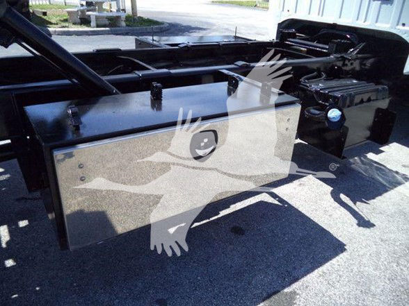 Truck chassis with large rectangular aluminum fuel/utility tank mounted under the frame; black components and blue cap visible on right; reflective surface shows surrounding area.