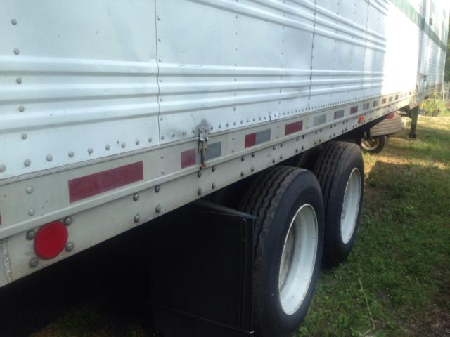 Trailer wheel area showing dual rear axles with tires and side skirt, corrugated metal trailer wall, reflective strips, and latch hardware.