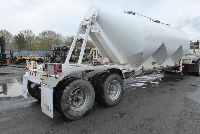 Truck trailer in a yard showing a large steel bulk grain hopper with three axles and dual rear tires. Visible details include a white painted body, rectangular tail lights, and support frame.