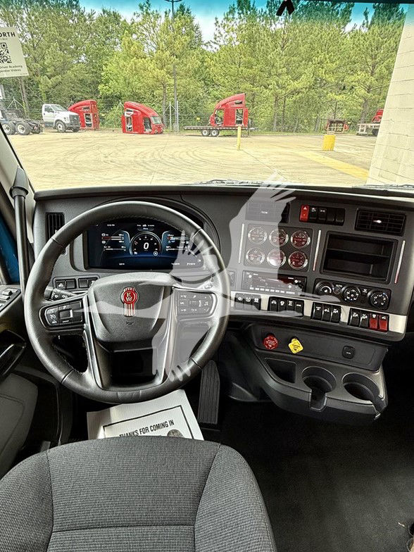 Kenworth T880 truck cab interior showing steering wheel, dashboard gauges, and controls; outside visible are red truck cabs and trees.