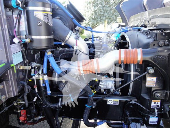Tow truck engine bay of a 2022 Peterbilt 337 showing blue hoses, turbo intercooler piping, black coolant reservoir, and various labeled stickers.