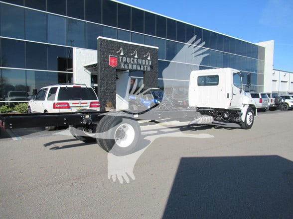 Truck chassis with cab and chassis at a dealership showroom. Visible text includes 'TRUCK WORLD KENWORTH' on a sign near the cab, and a large dark vertical plate with graphic elements. No other legible labels or numbers.