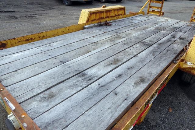 Flatbed trailer bed with weathered wooden planks and yellow metal rails, showing loading ramp at rear.