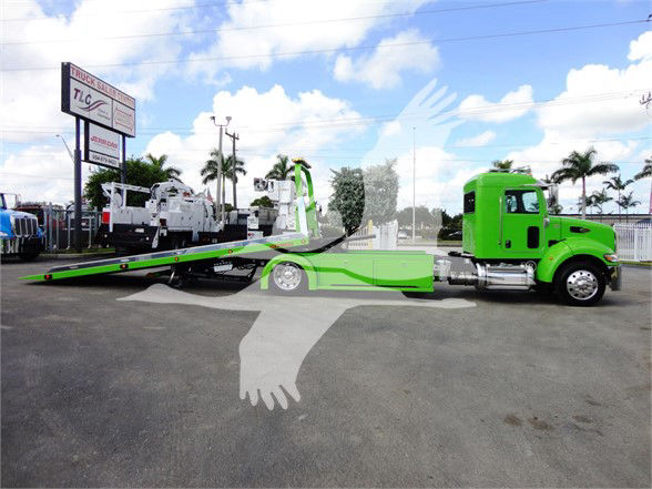 Green Peterbilt 337 tow truck with flatbed ramp in a dealership lot.