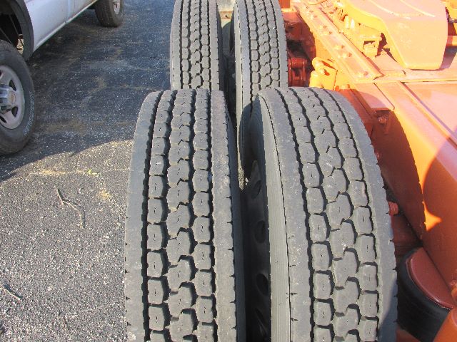 Truck rear dual tires mounted on a fifth wheel chassis with treads visible, orange cab in background, left side view.