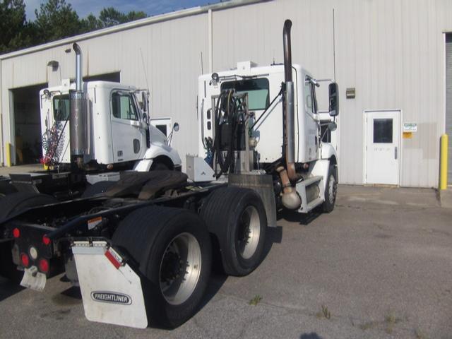 Front-and-side view of a white Freightliner Columbia conventional daycab tractor, showing dual rear axles and exhaust stacks; parking lot with a white industrial building in background.