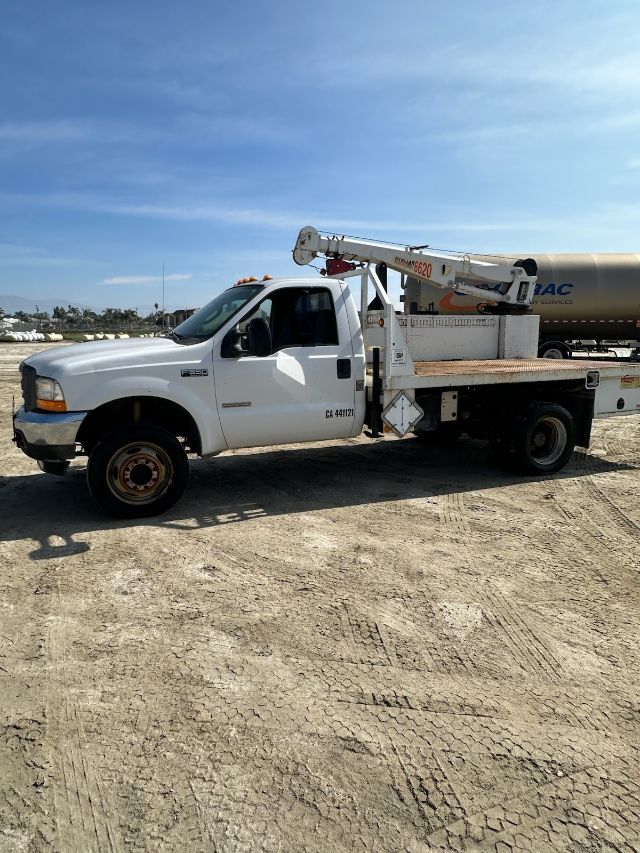 Flatbed tow truck with a mounted aerial lift, white cab, long bed, hydraulic boom folded over cab, California 44112 dock number, side warning decals visible.