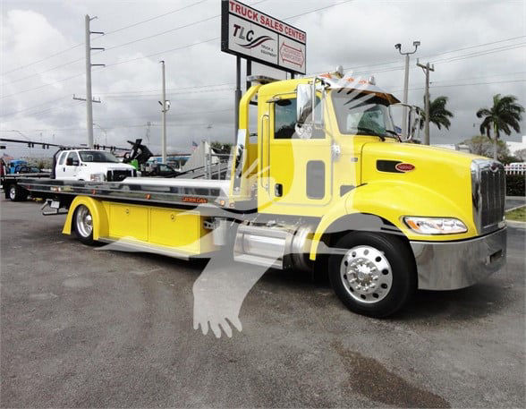Peterbilt 337 tow truck in yellow with flatbed, cab close-up showing chrome bumper and wheel, dealership lot in background.