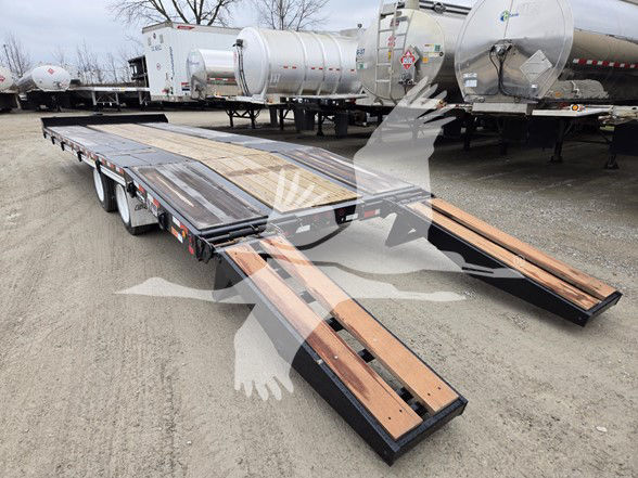 Trailer showing a long flatbed with folding ramps, wood decking in parts, black frame, and tandem axles with a white storage area in background.