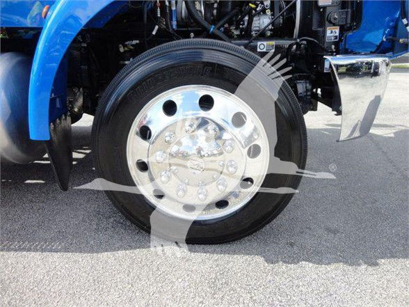 Wheel of a blue 2022 Peterbilt 337 truck showing polished aluminum hubcap and black tire, engine bay visible above, chrome front bumper on right.