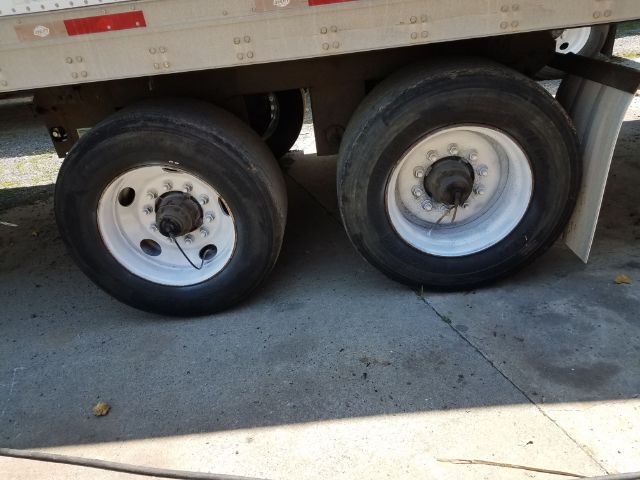Dual rear axles of a utility trailer showing two wheels with white rims and black tires, undercarriage with a metal side panel and mud flap.