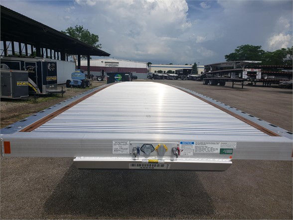 Flatbed aluminum trailer rear view showing ramp with labels and hitch, Fontaine Revolution 52 all aluminum 53ft tri-axle trailer text visible on tags.