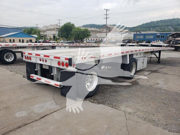 Flatbed trailer showing rear and side with taillights, reflective tape, and dual rear axles. Visible text includes brand decals along side rail.