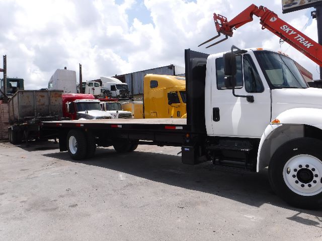 Flatbed tow truck with white cab and long flatbed, other trucks in background, forklift/crane arm visible