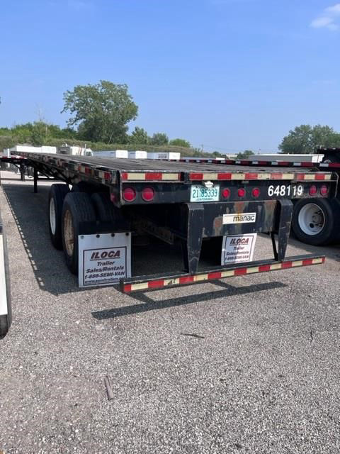 Manac flatbed trailer rear view showing dual axles, black frame, red/white reflective tape, and license plates.