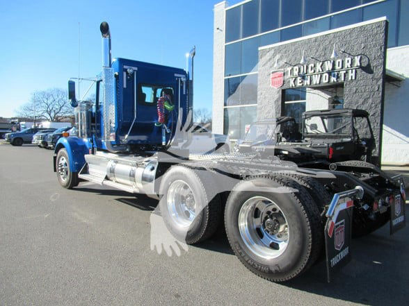 Kenworth W900L conventional daycab truck showing side profile with blue cab, chrome fuel tanks, rear dual wheels, and fifth wheel hitch.
