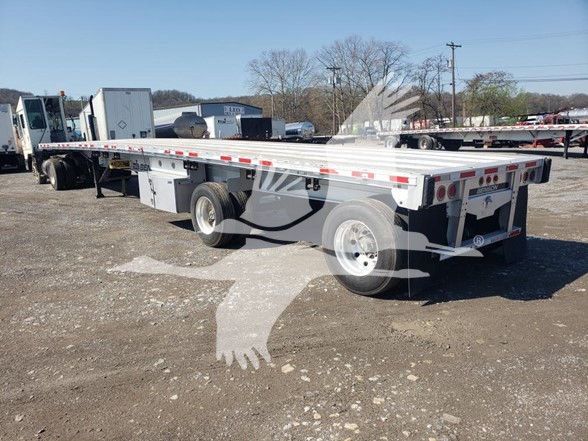 Flatbed trailer with long aluminum deck, dual rear axles, and white cab; badges and lights along sides; background shows other trailers and equipment.