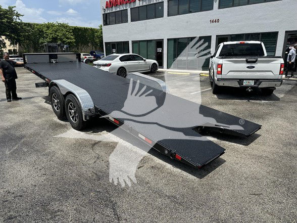 Flatbed trailer with dual drop ramps in a parking lot; background shows a white building with 'ADVANCED' signage and several parked cars; trailer has silver diamond plate surfaces and black deck.