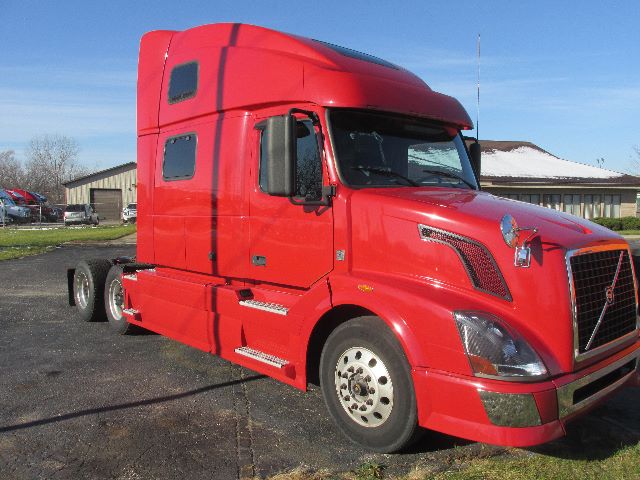 Volvo 780 conventional sleeper truck, red cab, side view showing front grille and wheel, daytime outdoor lot.