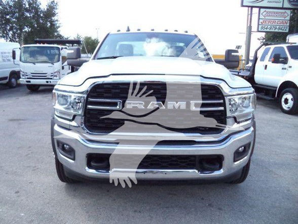 RAM front grille badge and chrome bumper of 2024 Ram 5500 SLT in a dealership lot with other white trucks in background.