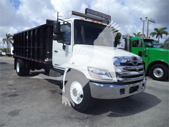 Truck in a dealership lot showing a white 2016 Hino 268A dump truck, with chrome grille, black dump bed, side mirrors, and a green truck in background.