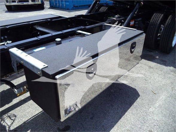 Truck chassis with a large rectangular toolbox mounted behind the cab. Toolbox has a textured black surface, with metal latches and a small circular port. Visible text includes 'M' and 'N' near the lower left corner of the toolbox.
