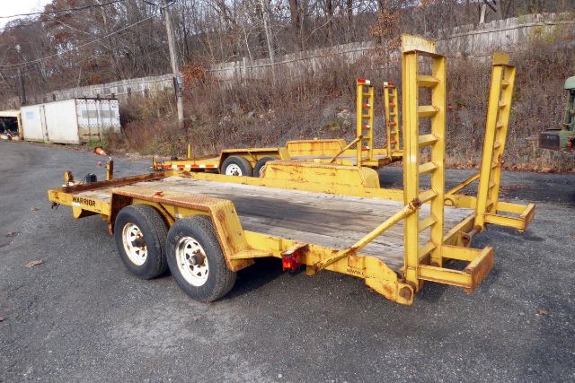 Military-style yellow heavy-duty trailer with dual rear axles, flatbed, and ramp/tie-down posts in an outdoor lot.