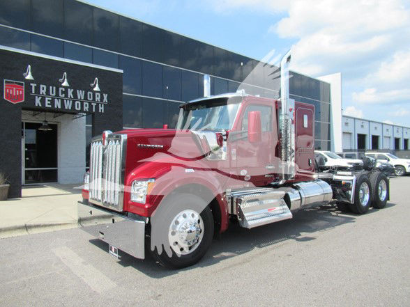 Kenworth W990 conventional sleeper tractor in red with chrome accents parked in front of a dealership showroom.