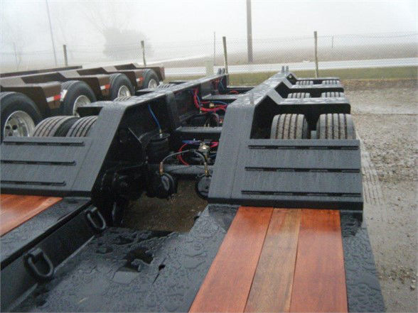 Lowboy trailer chassis with dual rear axles and hydraulic components visible between widened deck sections. Wet surface, wiring, and metal frame details.