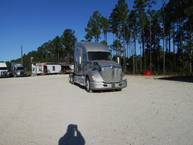Kenworth T680 cab and chassis truck in a gravel lot, silver/gray exterior, large chrome grille, trees in background.