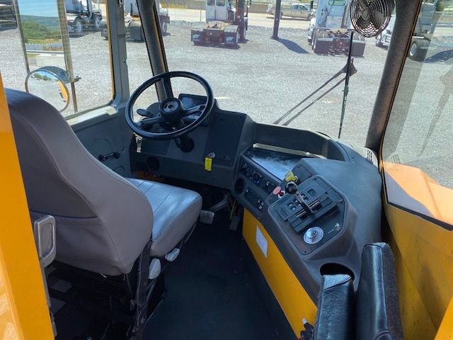 Operator cab of a used 2017 Ottawa T2 truck showing steering wheel, dashboard with controls, grey driver seat, yellow exterior, and windows with outdoor yard equipment in background.