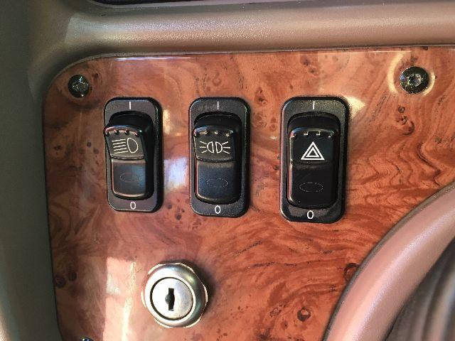 Panel inside truck showing three switches with labels; wood-grain bezel and ignition barrel above. Left switch triangle hazard symbol, middle switch with headlight icon, right switch with beam icon.