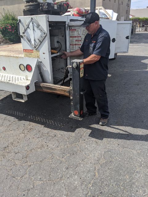 Truck lifting equipment with a man standing near the rear, rear of a white service truck, various labels and controls visible on the back.