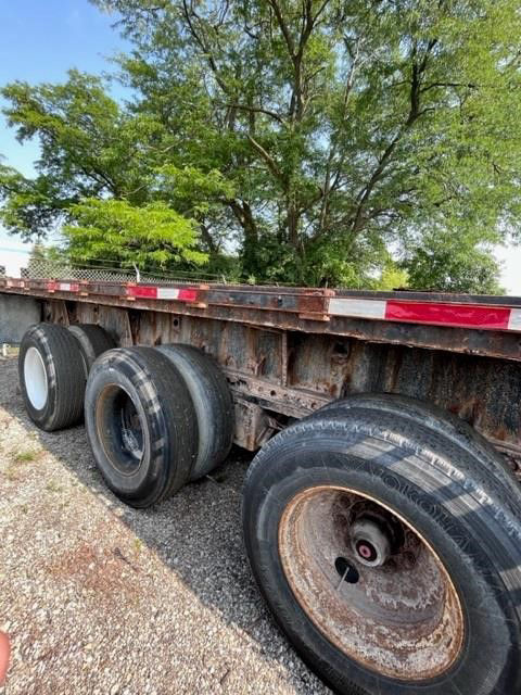 Flatbed trailer side with dual rear axles and rusted frame. Three tires visible on the near side with worn rims. Vegetation in background.