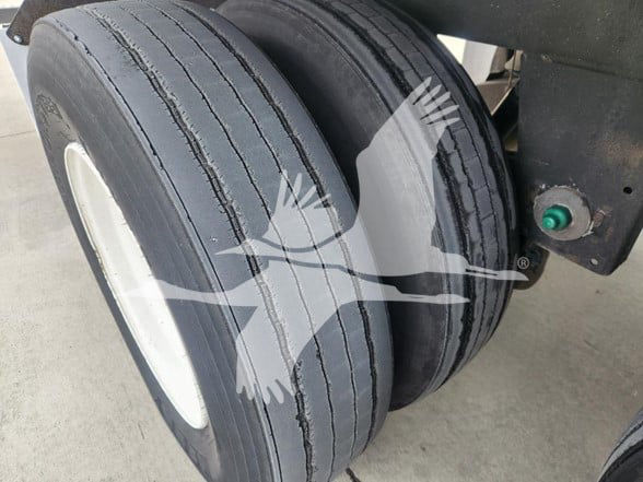 Dual rear trailer axles with black tires and white rims on a Wabash trailer, green sensor/bulb near frame, watermark of a bird logo overlaying image.