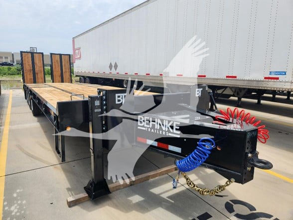 Flatbed trailer hitch showing Behnke Trailers branding, with blue coiled hose, black frame, wooden deck, and red safety chains.