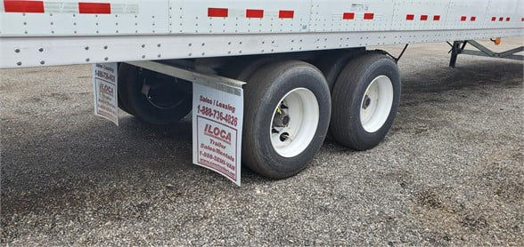Trailer rear wheels of a Vanguard VXP dry van with signs hanging from the mudflaps showing contact information and a stock number plate.