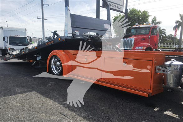 Tow truck with orange service body and a black flatbed, Peterbilt 337 in background. Visible text includes stock number J47166 and signage on the lot.