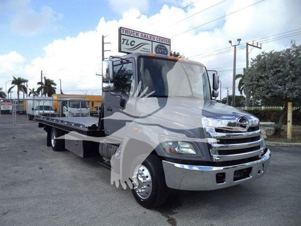 Tow truck cab with flatbed, Hino badge on grille, chrome bumper, daytime running lights, sign on top of cab.