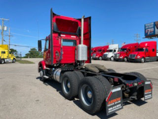 Volvo VNM64T-200 conventional daycab rear view showing chassis and dual rear axles, with red cab open doors, and multiple trucks in background.