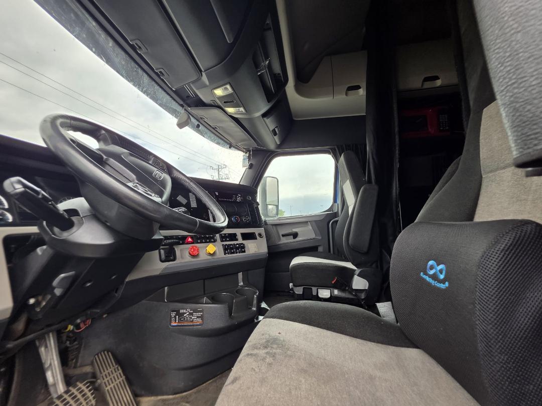 Freightliner Cascadia truck interior showing steering wheel, dashboard with controls, driver seat, and rack area.